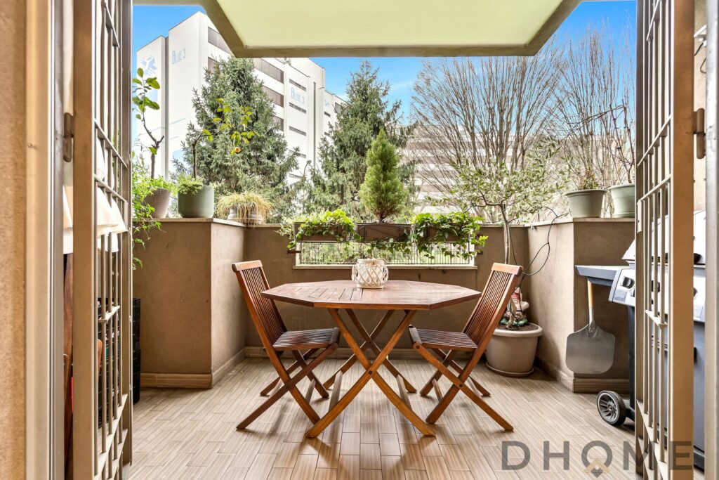 Balcone arredato con tavolo in legno e piante verdi, vista su edifici e alberi in città.