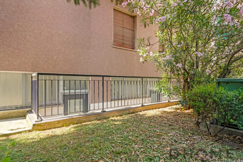 Balcone esterno con ringhiera in ferro e barbecue, vista su giardino con alberi e piante.