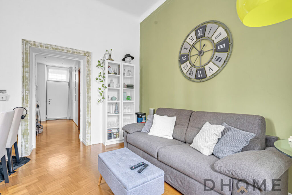 Soggiorno moderno con parete verde, grande orologio e divano grigio, arredamento elegante e accogliente.