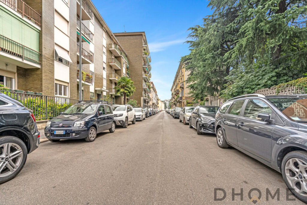 Auto in sosta lungo strada residenziale a Milano, appartamenti con balconi e piante, cielo sereno, ambiente urbano tranquillo.