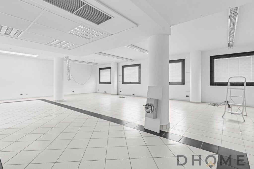 Ampia stanza ufficio in costruzione, pareti bianche, finestre grandi, ambiente luminoso.