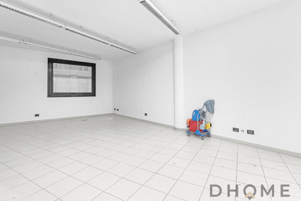 Spazio ufficio vuoto con pareti bianche e pavimento in ceramica, illuminato da luci a soffitto e finestra grande.