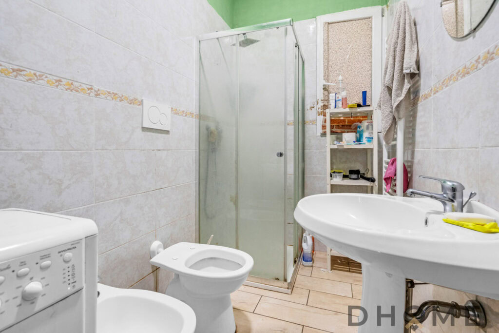 Bagno piccolo con asta aldo in vetro e sanitari bianchi, pavimento in piastrelle beige e pareti chiare.