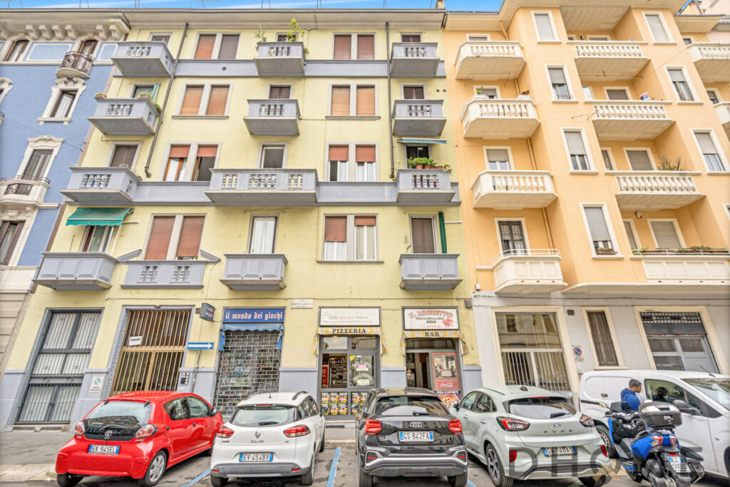 Colorful residential building with commercial spaces on ground floor, parked cars, and street scene in an Italian city.