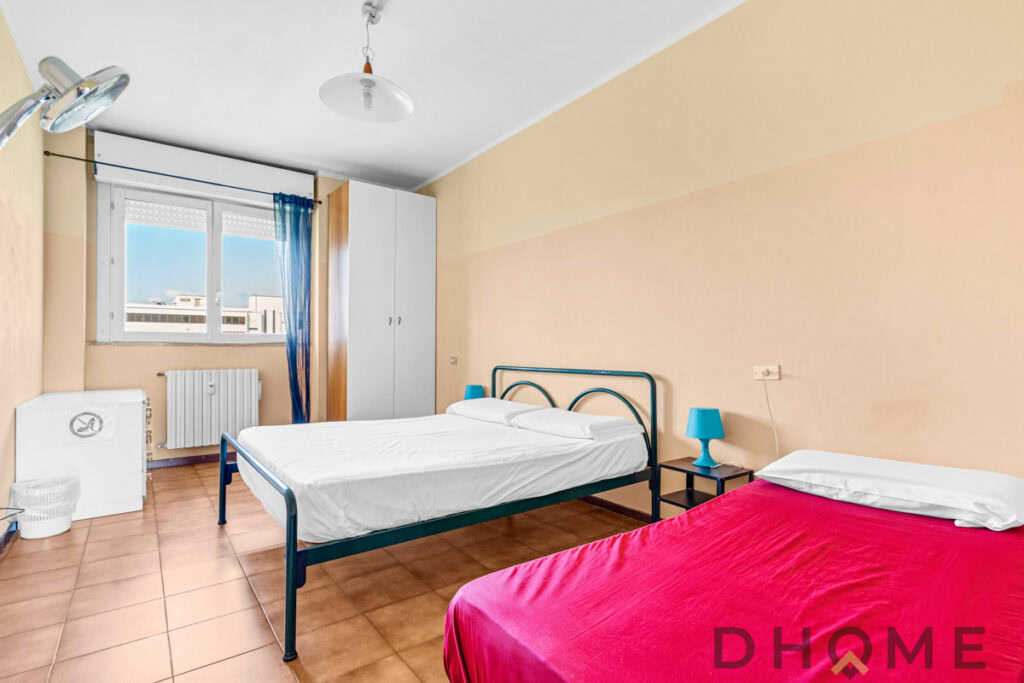 Letto singolo con biancheria in camera doppia, arredamento semplice, finestra con tende blu, atmosfera luminosa e confortevole.