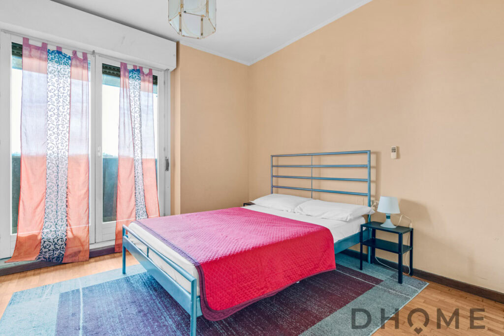 Letto matrimoniale in camera con pareti beige e tessuto rosso sulla coperta, con tende colorate sulle finestre.