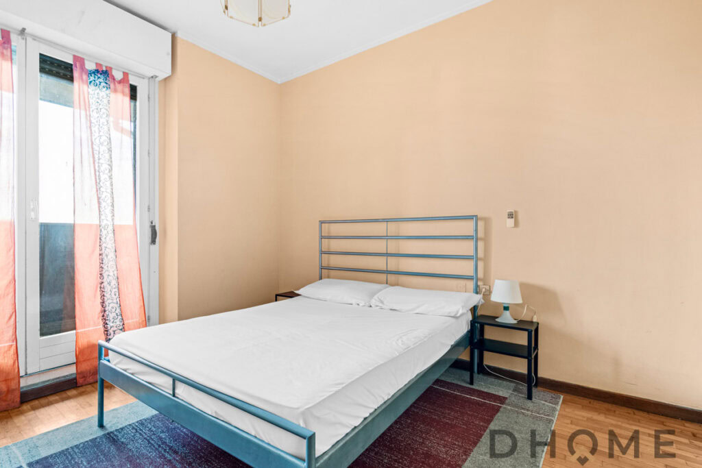 Letto matrimoniale in camera con arredi semplici e luminose tende da sole.