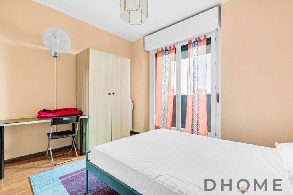 Letto matrimoniale in camera con balcone e tende colorate, ambiente estivo e luminoso.