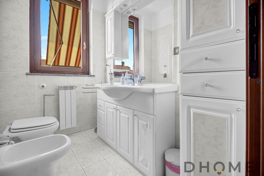 Lavandino e mobili bagno bianco in stile classico, finestra con vista su tetti e cielo, bagno luminoso e funzionale.