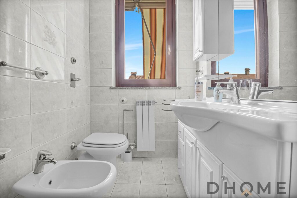 Lavabo bianco con mobile bagno e finestra con vista su tetti e cielo azzurro, esterno luminoso.