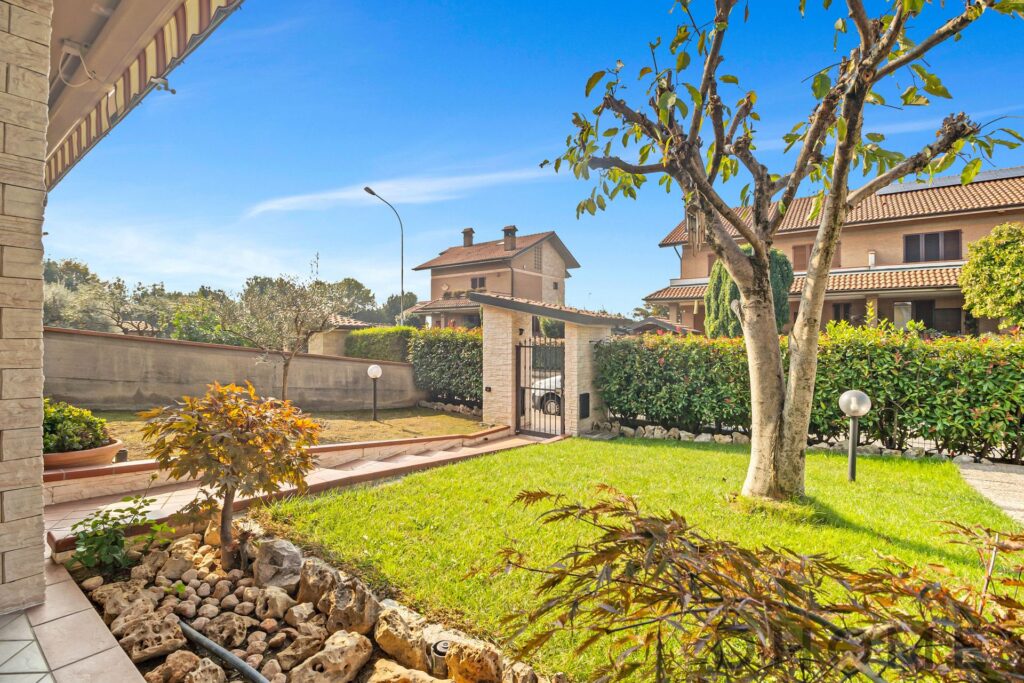 Amaca giardino con prato verde, alberi e siepi, vista di case residenziali, ambiente tranquillo e soleggiato.