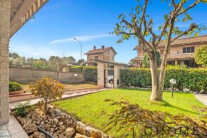 Amaca giardino con prato verde, alberi e siepi, vista di case residenziali, ambiente tranquillo e soleggiato.