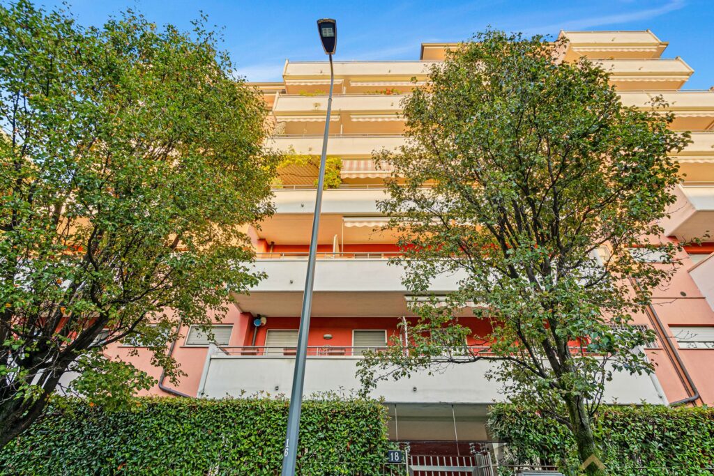 Balcone di un appartamento moderno con alberi e cielo azzurro, facciata colorata e arredi eleganti, sito in zona residenziale.