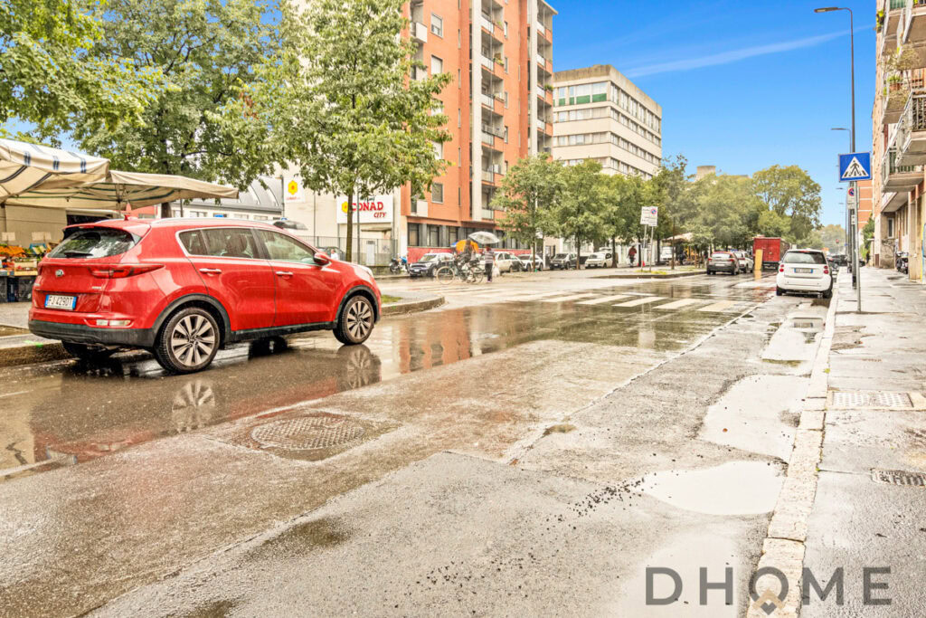 Auto rosso in via urbana sotto la pioggia, edifici residenziali e negozi in background, strada molle e bagnata.