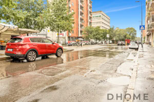 Auto rosso in via urbana sotto la pioggia, edifici residenziali e negozi in background, strada molle e bagnata.