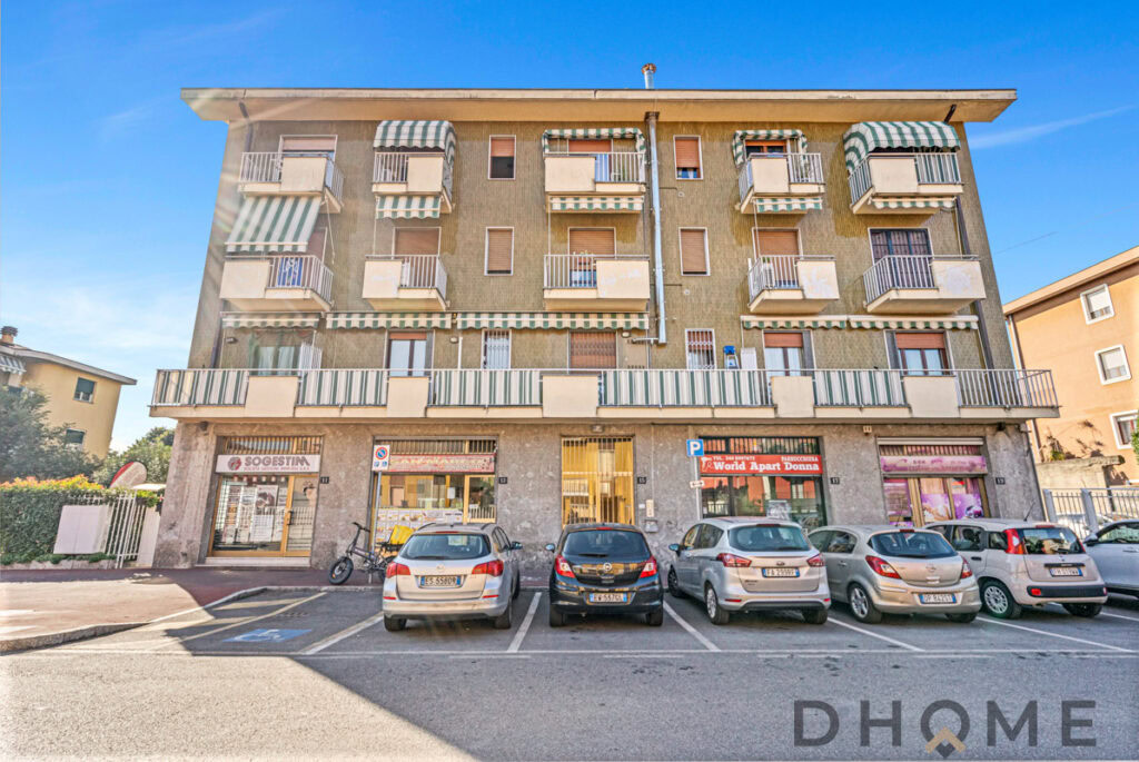 Appartamenti con balconi e negozi al piano terra in edificio residenziale, parcheggio esterno.